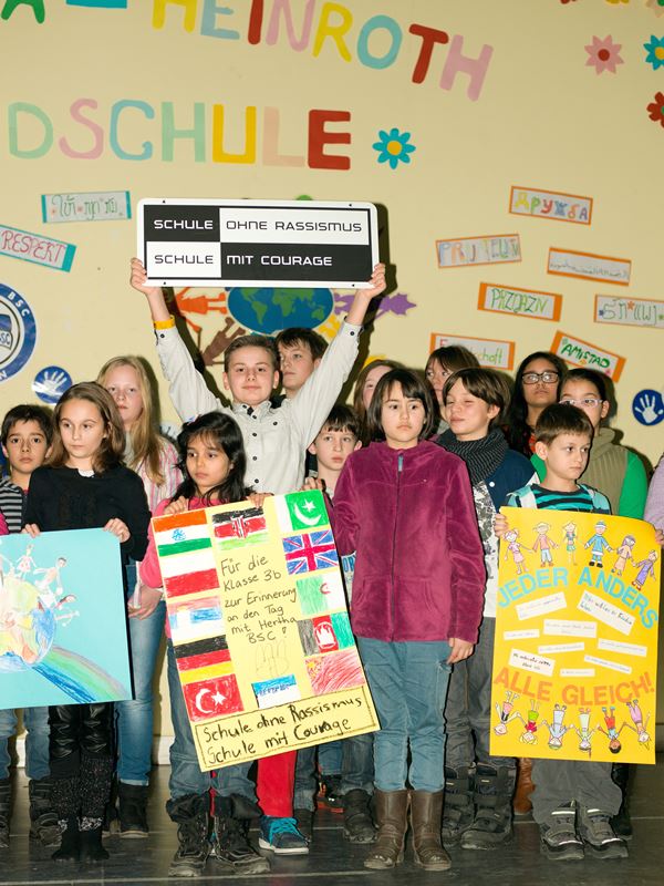 Vierzehn Kinder stehen auf der Bühne in der Aula, drei halten stolz Plakate in der Hand zum Thema Anders sein sowie eines mit der Aufschrift "Schule ohne Rassismus" in den Händen