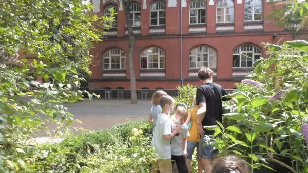 Kinder ernten im Garten mit einem P&auml;dagogen, im Hintergrund ein rotes Backsteinschulgeb&auml;ude