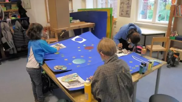 Im Werkraum kleben Kinder in Kitteln Elemente auf gro&szlig;e blaue Pappen