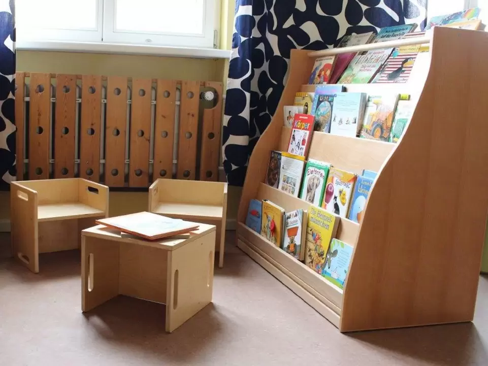 Leseregal rechts mit B&uuml;chern, links steht ein kleiner Tisch mit zwei kleinen Sesseln aus Holz