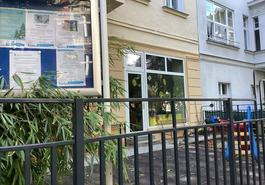 kleines Gitter, dahinter der Eingang zu der Kita mit bunter Fensterfläche, links ein Schaukasten