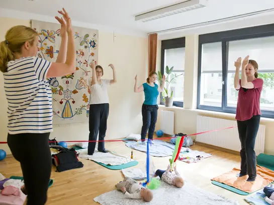 Vier Frauen stehen im Gymnastikraum auf Matten und machen &Uuml;bungen der R&uuml;ckbildungsgymnastik. Die Kinder liegen auf Decken am Boden und spielen mit aufgeh&auml;ngten T&uuml;chern.