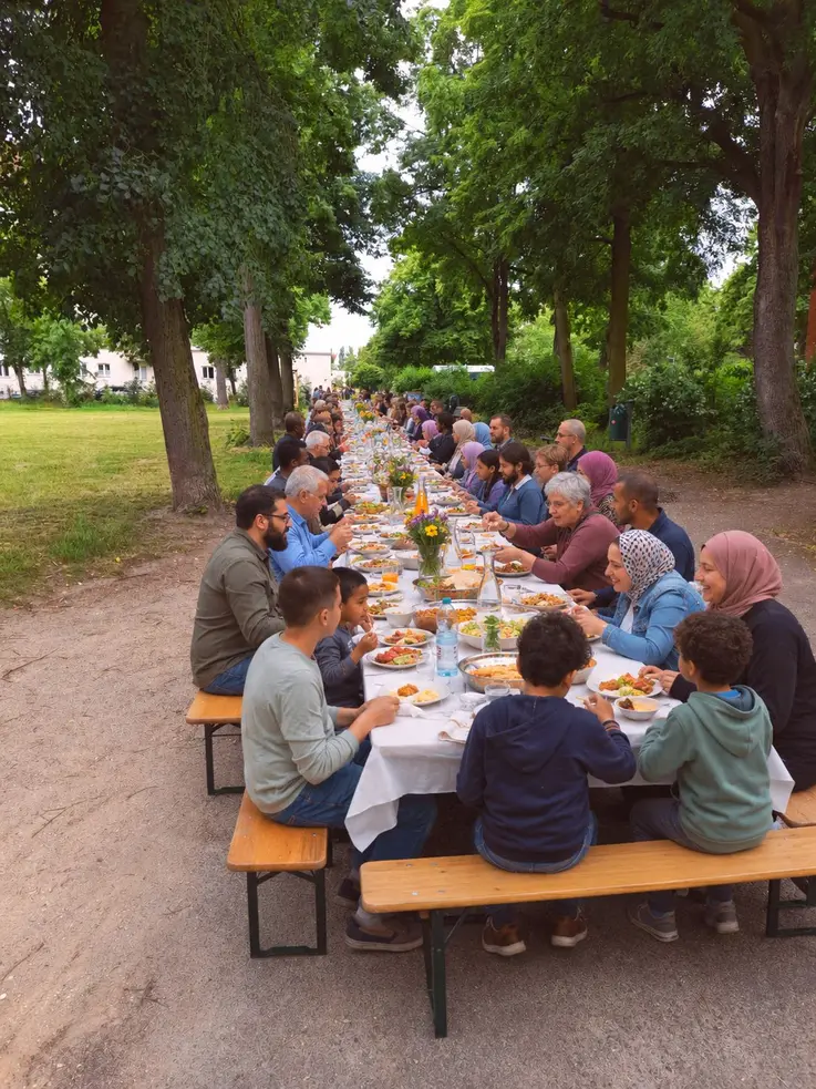 Auf einem Platz, zwischen B&auml;umen und Wiese, sitzen an einer langen Tafel viele alte und junge Menschen und essen gemeinsam.