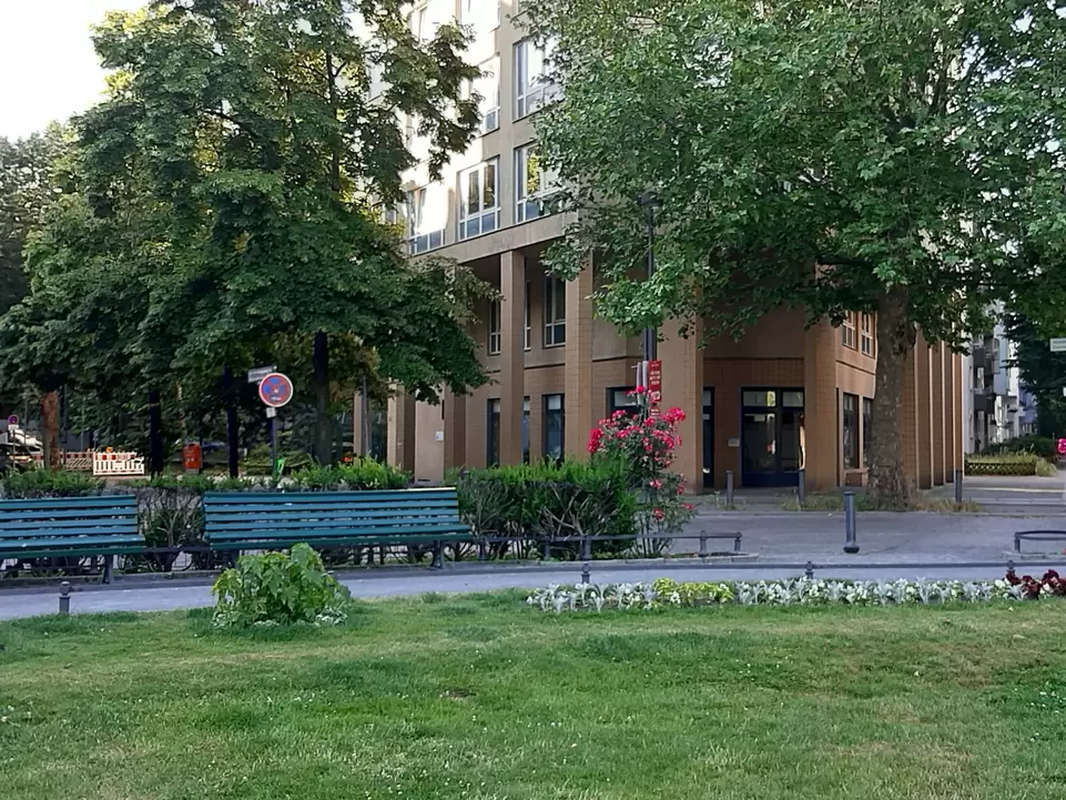 Platz mit Wiese und Parkb&auml;nken, dahinter ein hellbraunces Geb&auml;ude