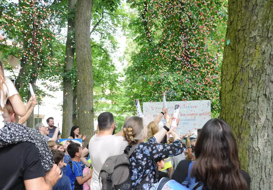 Familien mit Kindern zwischen B&auml;umen stehen vor einem Plakat, buntes Konfetti regnet herab