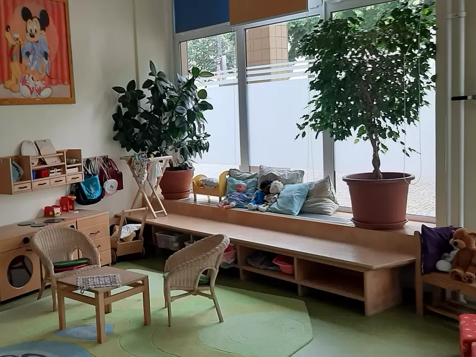 Raum mit kleinem Tisch und Sesseln, rechts eine Holzbank mit Kissen und zwei gro&szlig;en Pflanzen. Puppen und Spielsachen im Regal.