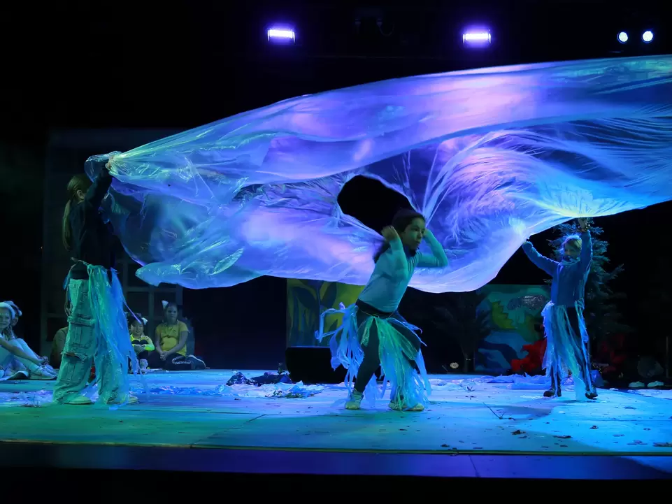 Kinder stehen in Kost&uuml;men auf der B&uuml;hne und wirbeln ein gro&szlig;es Tuch, dass lila-gr&uuml;n erleuchtet wird